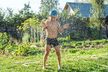 Joyful boy in a cap enjoys splashing water from a hose, playfully cleaning himself in a vibrant garden under bright sunshine