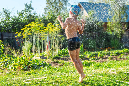 Joyful boy in a cap enjoys splashing water from a hose, playfully cleaning himself in a vibrant garden under bright sunshine