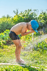 Joyful boy in a cap enjoys splashing water from a hose, playfully cleaning himself in a vibrant garden under bright sunshine