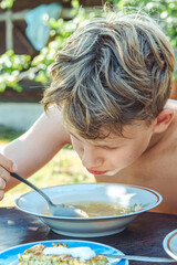 Boy happily eats his lunch from a bowl while sitting at a wooden table in a bright, sunny garden of a countryside home, closeup