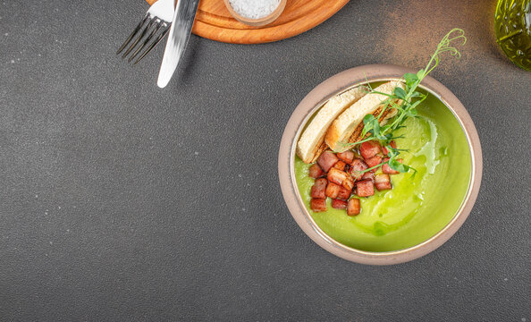 Bowl of creamy green pea soup garnished with crispy bacon cubes, fresh herbs, and toasted bread slices, served on a dark textured background - Powered by Adobe