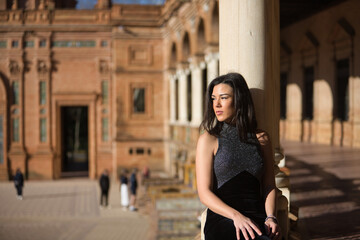 Portrait of young, beautiful, brunette woman in a fancy black dress, sitting and leaning on a marble column, looking at the square of Spain in Seville. Concept beauty, trendy, femininity, tourism.
