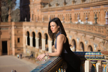 Young, beautiful, brunette woman in a fancy black dress, leaning on a railing on a balcony overlooking the square of Spain in Seville. Concept beauty, fashion, trendy, femininity, tourism.