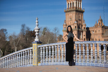 Young, beautiful, brunette woman in a fancy black dress, posing looking at the camera, leaning on a tiled railing on a bridge in the square of Spain in Seville. Concept beauty, femininity, tourism.