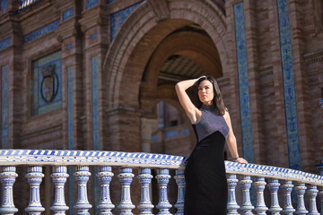 Young, beautiful, brunette woman in a fancy black dress, looking at infinity, leaning on a tiled railing on a bridge in the square of Spain in Seville. Concept beauty, fashion, femininity, tourism.