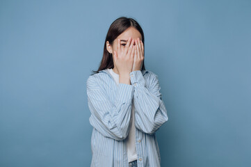 Young woman covers her face with hands, expressing emotions in front of a blue background during a...