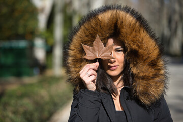 Portrait of a young, beautiful, dark-haired woman in a fur hooded coat, covering her eye with a dry leaf, on a cold, sunny day. Concept winter, cold, autumn, weather.