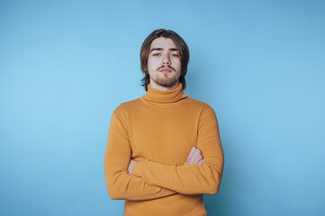 Confident young man in yellow turtleneck sweater stands against blue background with arms crossed...