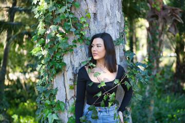 A young, beautiful, dark-haired woman in a black T-shirt and jeans, leaning on the trunk of a tree and entangled in ivy, in connection with nature. Concept beauty, femininity, nature, plants, trees.