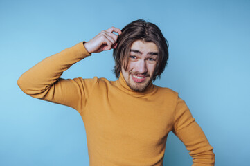 Young man scratching his head and smiling against a blue background while wearing a yellow sweater