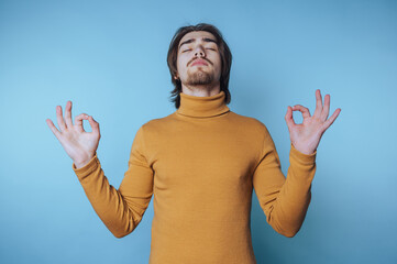 Young man meditating calmly against a light blue background while wearing a cozy turtleneck sweater