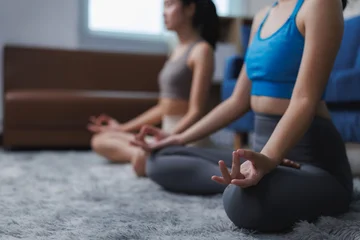  Two young women meditating together in lotus position at home © Phimwilai