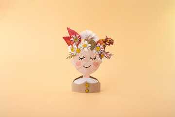 Cardboard character with floral headpiece on pastel peach surface, ideal for eco toy branding,...