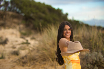 Young, pretty, brunette woman, in a yellow dress, offering a hat with her hand, looking at the...