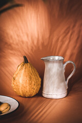 A charming still life captures autumn vibes with a gourd and pitcher. A still life composition features a gourd and a white pitcher against a warm-toned background, with soft shadows.