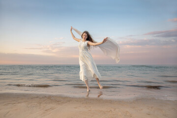 A young woman in a flowing white dress dances freely with a shawl by the sea at sunset. A joyful, dreamy scene with soft light and a pastel sky.
