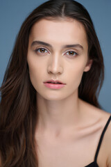 Obraz premium Close-Up Portrait of a Young Woman with Brown Hair and Delicate Features Against a Soft Blue Backdrop Showcasing Natural Beauty and Youthful Radiance for Skincare and Cosmetics