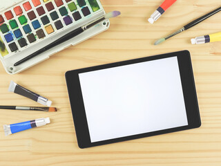 flat lay of artist workspace with digital tablet. Drawing tools, stationary supplies, Watercolor paints, brushes and white blank screen of digital tablet on wooden table, copy space.