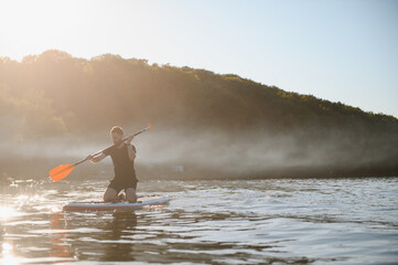 Man kneeling on sup board paddling on misty lake at sunrise