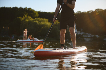 Sportsman paddling on sup board at sunset on lake with pedal boat in background