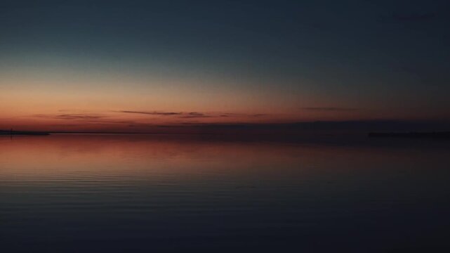 A peaceful sunset over the calm sea. Vibrant orange and blue gradient sky reflects on still water. Minimalist seascape filmed at twilight, ideal for background or meditative footage.
