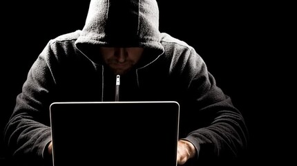 A hooded figure intently working on a laptop, shrouded in shadow.