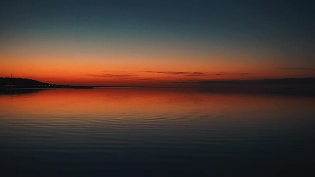 A peaceful sunset over the calm sea. Vibrant orange and blue gradient sky reflects on still water. Minimalist seascape filmed at twilight, ideal for background or meditative footage.