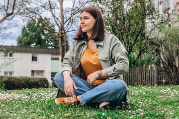 Young woman sits on grass, smelling a flower on a sunny day. Calm moment in nature, symbolizing...