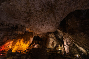 Visitors explore a vast cave featuring impressive stalactites and stalagmites, enhanced by vibrant lighting that highlights the natural formations. The atmosphere is serene and mysterious