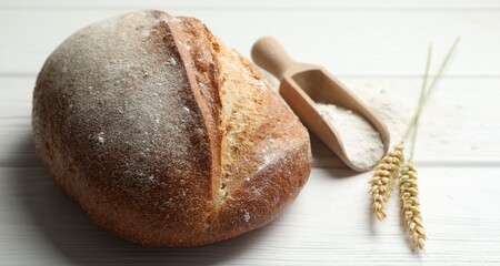 Loaf of fresh bread, flour and spikes on white wooden table, closeup