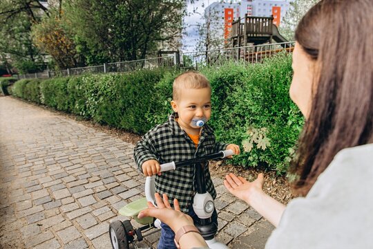 2 year old toddler rides bicycle joyfully toward mother open arms outdoors. A loving moment of bonding, play, learning, and family connection in a sunny park