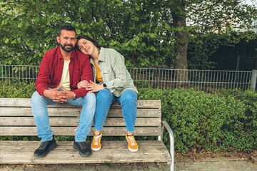 Happy couple sits on a bench outdoors, smiling and hugging each other. A joyful, affectionate...