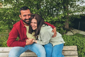 Happy couple laughs as boyfriend tickles girlfriend on park bench. Joyful moment of affection,...