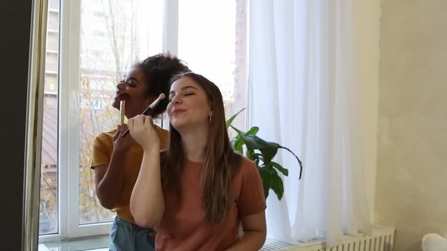 	187 charsTwo young women applying makeup in a bright room with large windows, showcasing a fun and engaging beauty routine with plants in the background, emphasizing friendship and self-expression
