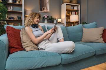 Obraz premium Young woman or girl sitting at home on sofa holding digital tablet, searching for music to play on wireless headphones. Comfortable happy female choosing streaming service for fun or online dating.