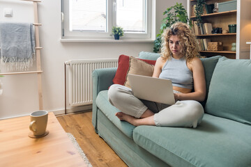 Concentrated millennial girl sit on couch working on laptop browsing internet at home during lazy...