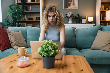 Concentrated millennial girl sit on couch working on laptop browsing internet at home during lazy weekend, focused young woman freelancer busy using computer surfing wireless web shopping online