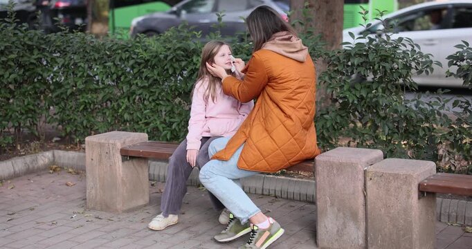 Woman wipes daughter nose with handkerchief sitting near mom on bench outdoors. Mother helps little girl to blow runny nose in city park slow motion