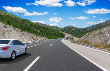 A white sedan drives along a winding mountain highway under a bright blue sky with scattered...