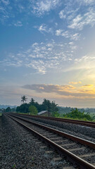 Obraz premium A peaceful railway track passing through the countryside with rice fields, tropical trees, and a warm sunrise in the background.