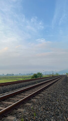 Fototapeta premium A railway track running through vast rice fields with misty morning sky and tropical greenery in a rural countryside setting.