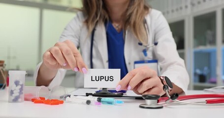 Female doctor in lab coat holds card with word Lupus at desk. Instruments and pill bottles indicate clinical setting focused on medical conditions