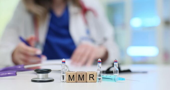 Lady medical professional reviews documents near MMR blocks on desk. Numerous vials syringe and stethoscope indicate focus on MMR vaccine in hospital