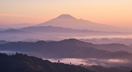 Landscape of majestic volcanic mountain peak rising above misty purple and orange layers of hills at sunrise