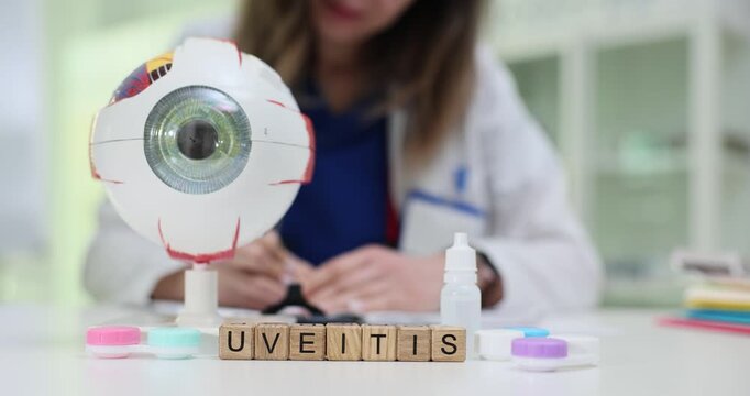 Cubes form word Uveitis placed under giant eye model on table. Each wooden tile stands upright showing full name while medical bottles lie nearby