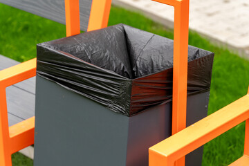 Trash bin with black liner and bright orange frame sits near a park bench, emphasizing urban public...