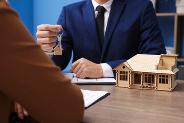 Real estate agent giving key to client at desk in office, focus on house model