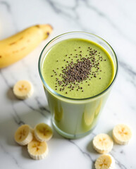 Top view of refreshing vegan smoothie made with matcha and maca in transparent glass, placed on marble surface with banana slices and chia seeds, clean white backdrop
