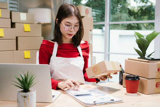 Young Asian woman in red shirt and apron running her online business, multitasking between smartphone and laptop, surrounded by packaging boxes in modern workspace.