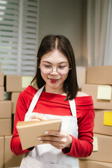 Young Asian woman in red shirt and apron doing online business, multitasking between smartphone and laptop, surrounded by packaging boxes in modern workspace.